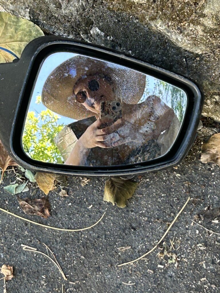 Photo d’une femme souriant dans le reflet d’un rétro situé au sol, illustrant la page À propos d’Emilie Barbe.